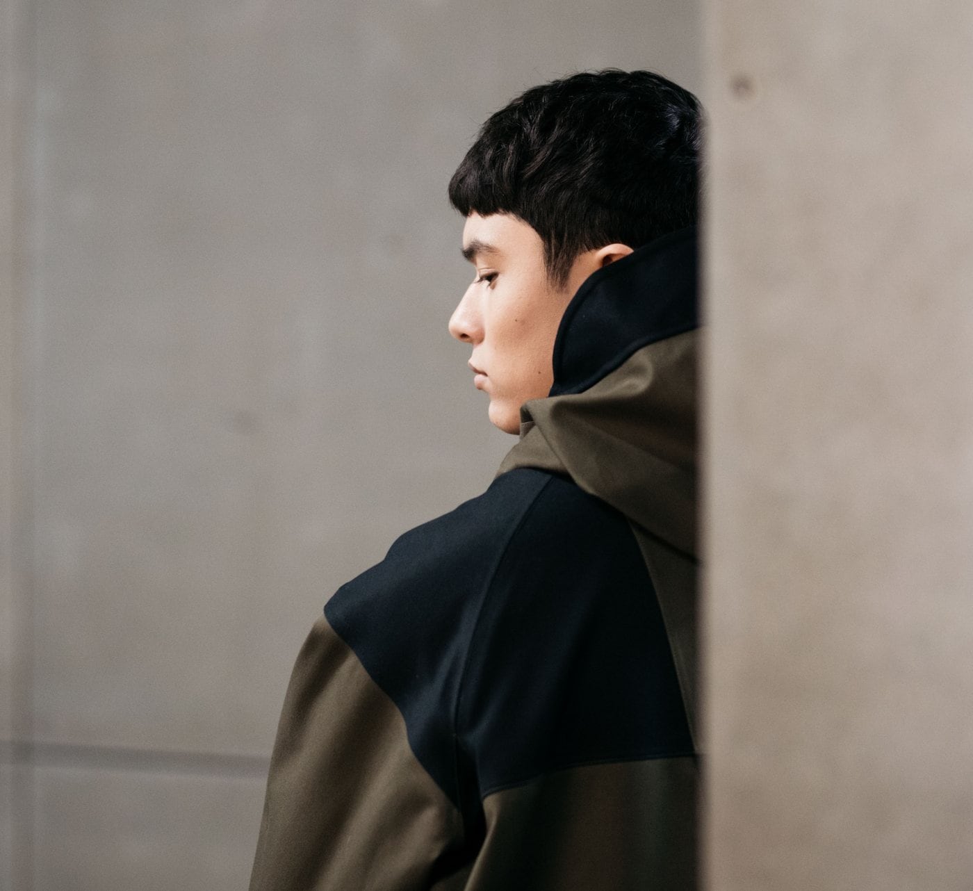 A man in a dark hooded jacket, in profile beside a concrete wall